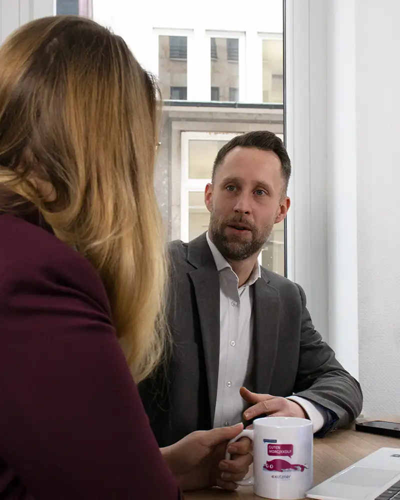 A man with a short beard and a friendly expression in a suit and a woman sit at a table. The two are engrossed in a consultation.
