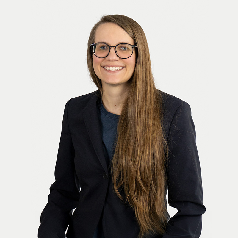 A woman in a black blazer with a blue shirt, nerd glasses, and long brown-blonde hair. She smiles at the camera. 