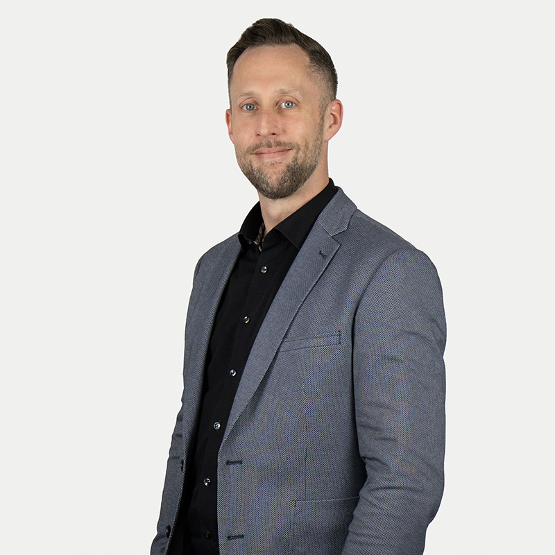 A man in a gray casual jacket and black shirt. He has short dark-blond hair and a short beard and looks friendly into the camera.